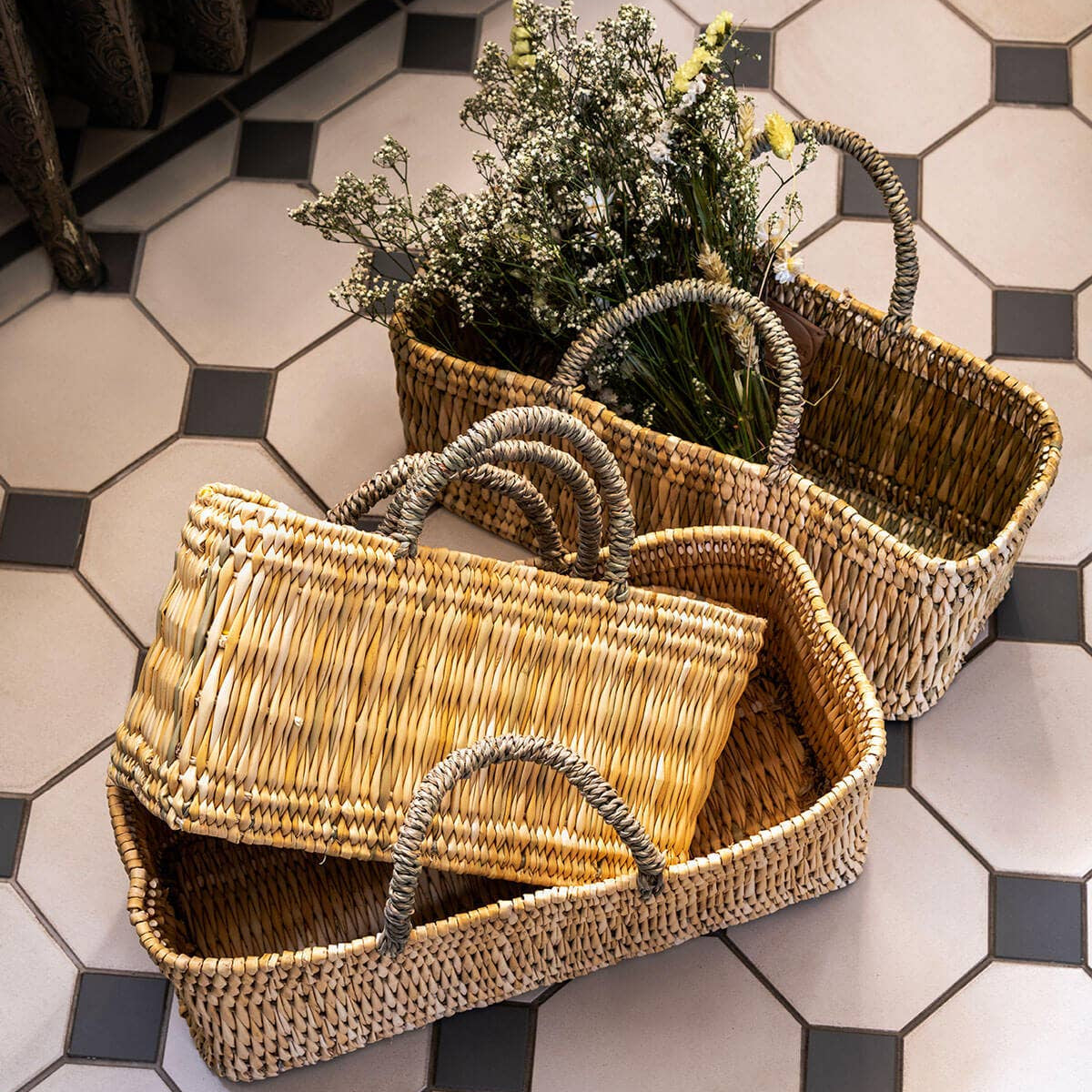 Traditional Moroccan reed baskets with sisal handles, placed on a tiled floor. One basket contains flowers, and the others are empty.