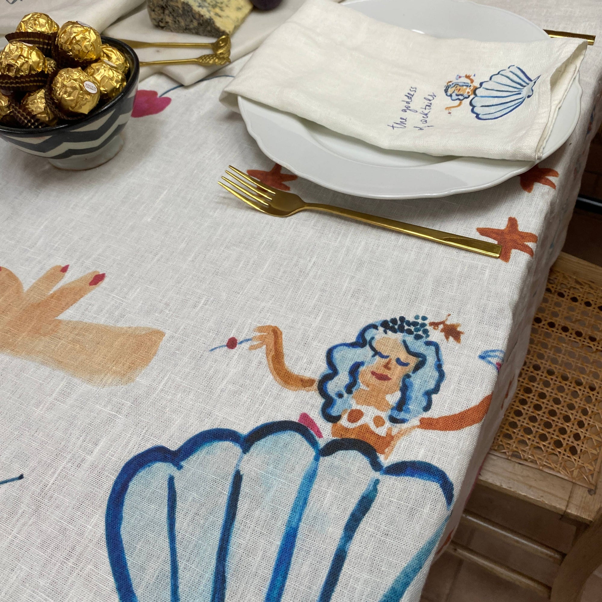 Table setting with a decorative tablecloth featuring mermaid and shell design, plates, and gold cutlery.