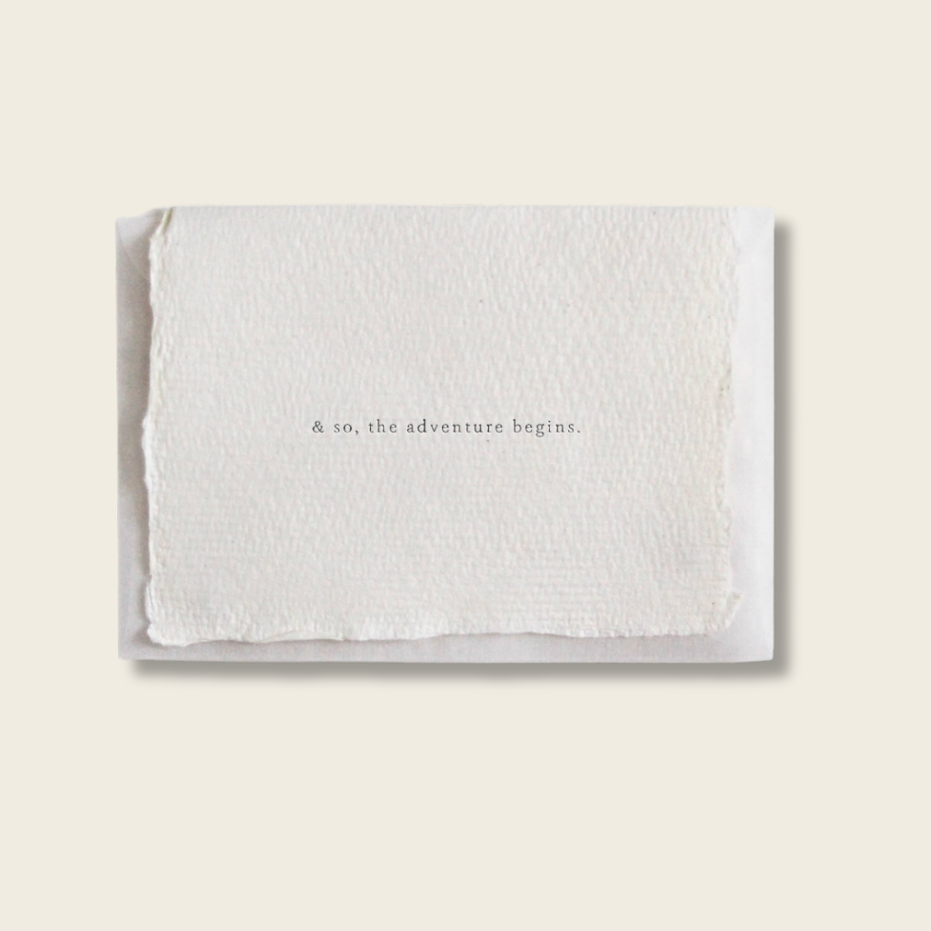 White folded paper with 'and so, the adventure begins' text on a light beige background