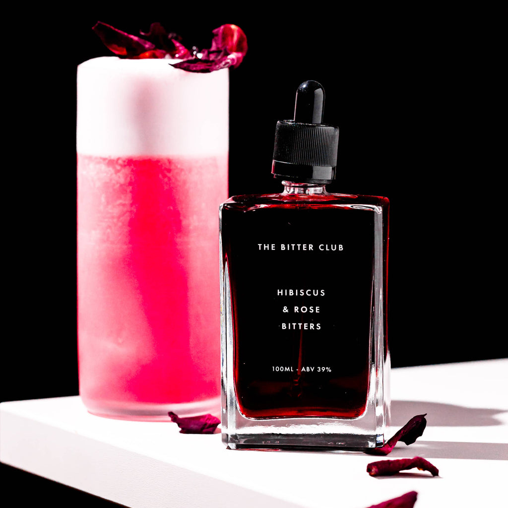 Bitters bottle labeled 'The Bitter Club' next to a pink cocktail with a black background