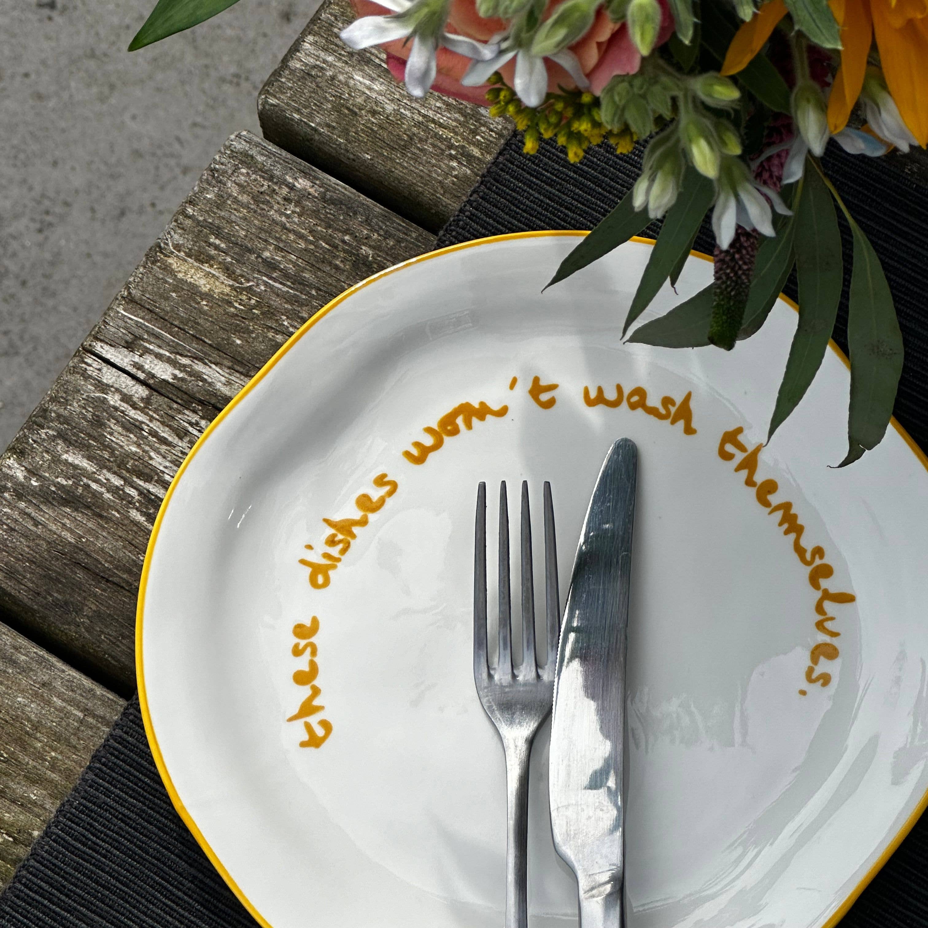 Plate with gold text, fork, knife, and colorful flowers on a wooden surface