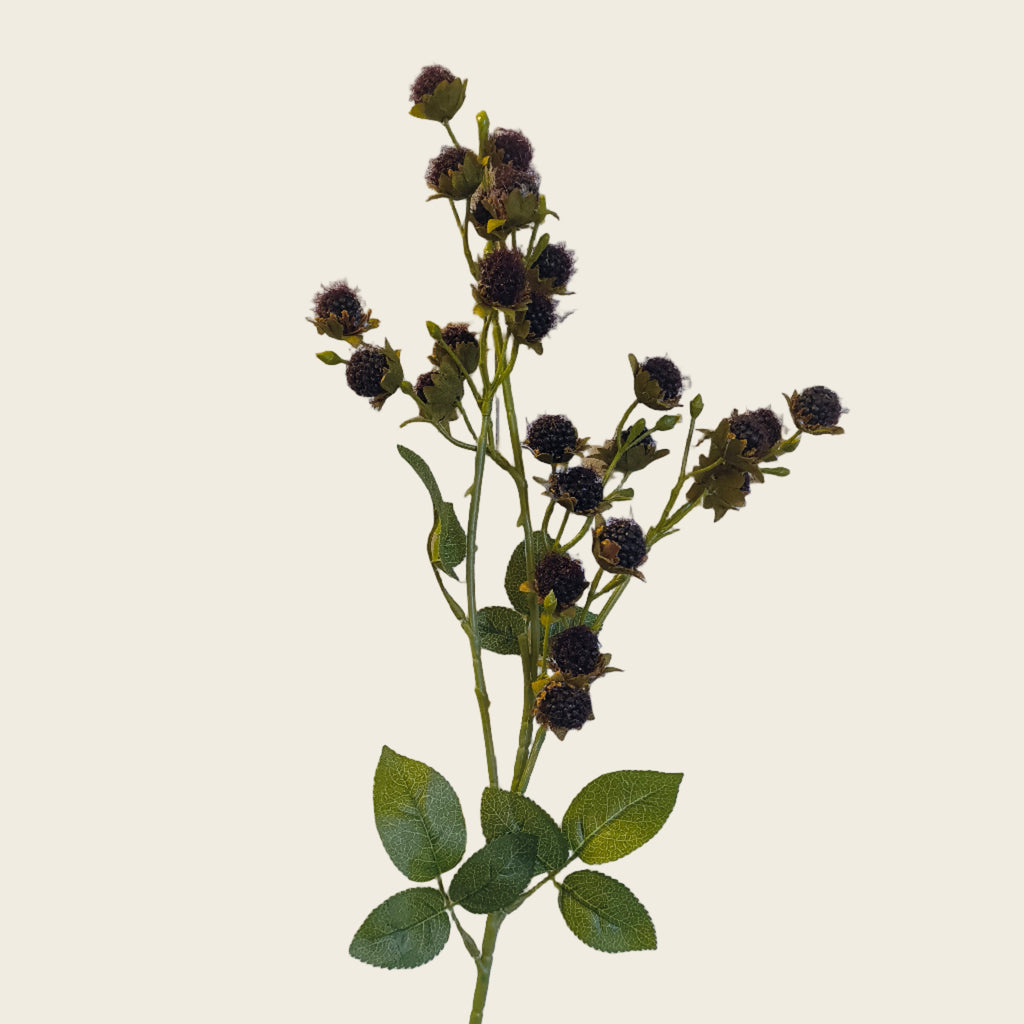 Artificial flower stem with dark buds and green leaves against a light wooden panel background