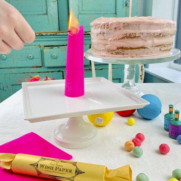 Pink candle being lit on a cake stand with a cake and colorful balls on a table.